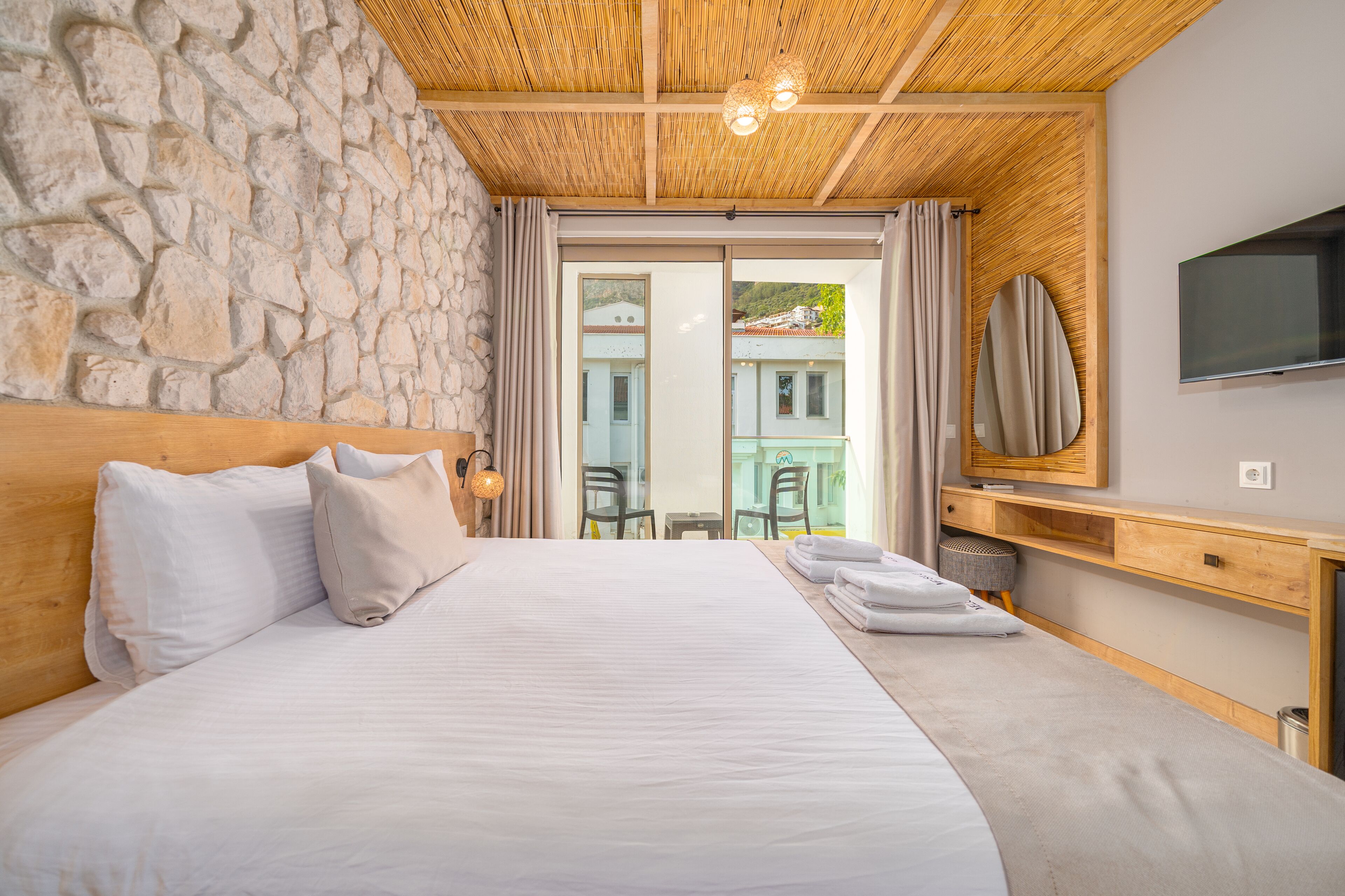 A bedroom with a stone wall, a wooden ceiling, a bed with pillows, a TV, and a view of the outside.