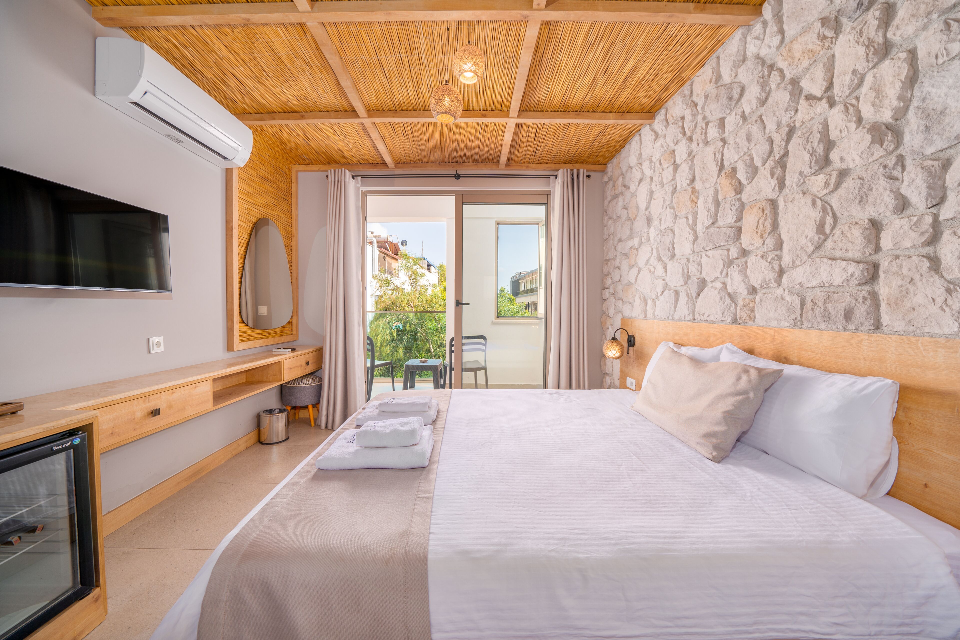 A modern hotel room with a large bed, a flat-screen TV, a wooden desk, and a stone wall.