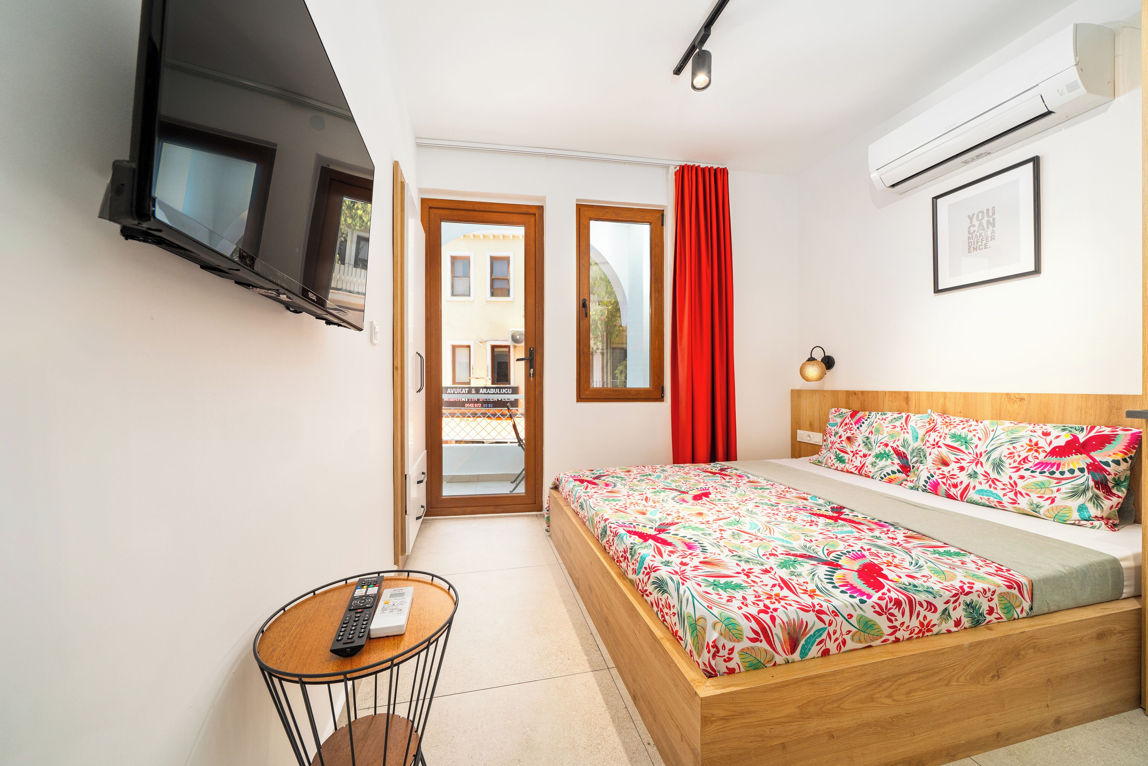 A bedroom with a bed, a TV mounted on the wall, a small table with a remote, and a door leading outside.