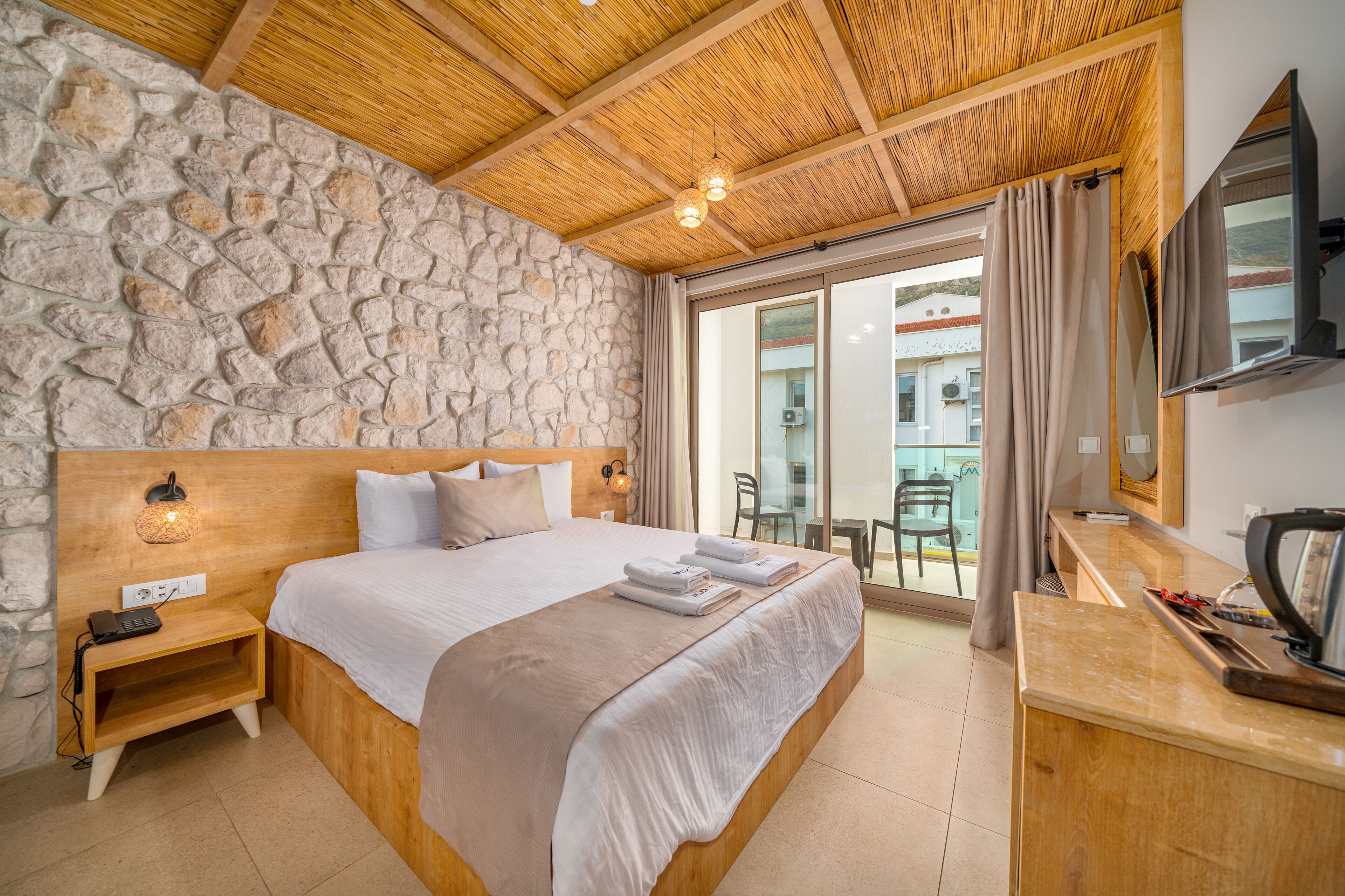 A bedroom with a stone wall, wooden ceiling, a bed with white linens, a bedside table with a lamp, and a TV on the wall.