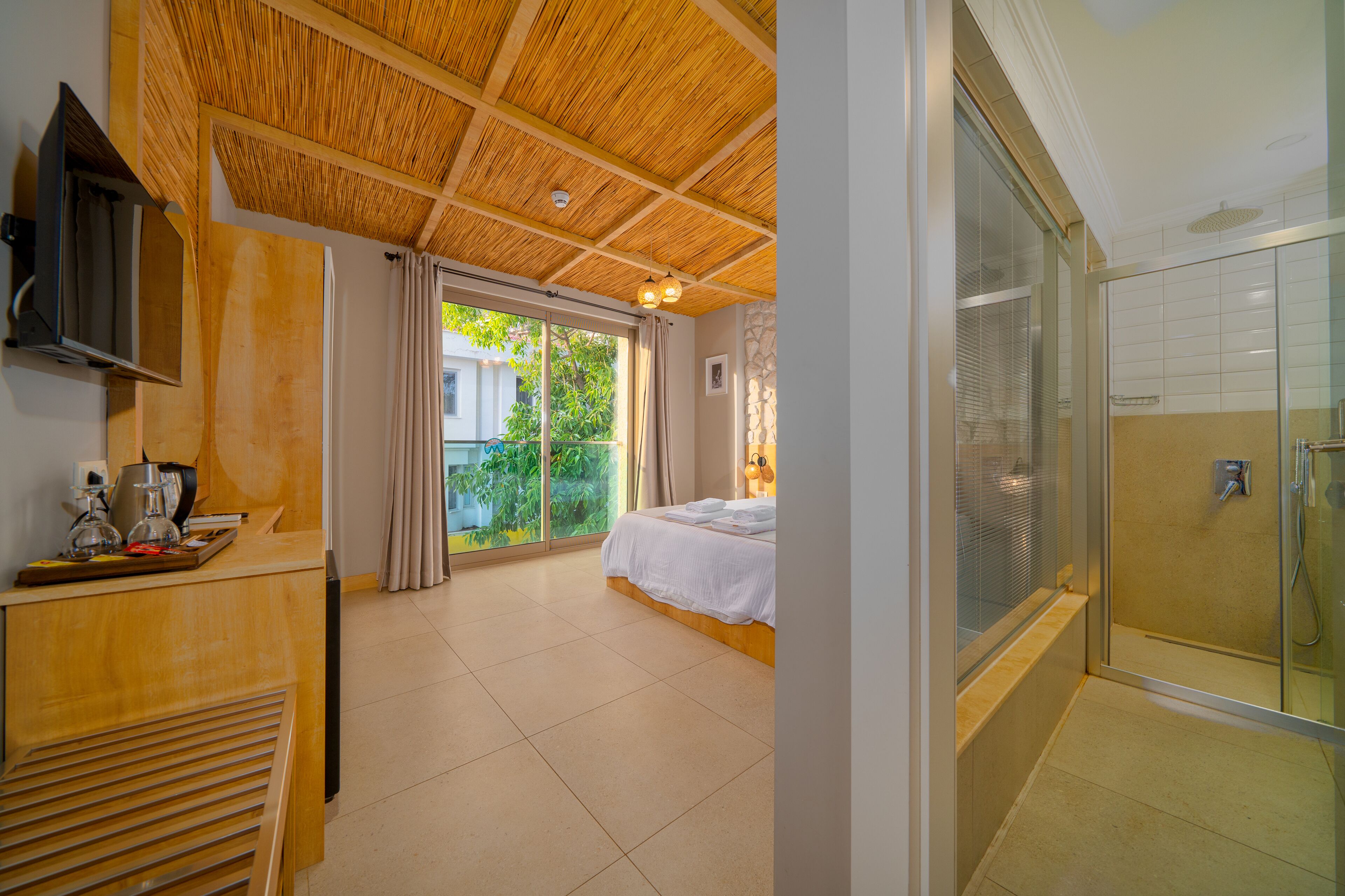 Quarto de hotel com teto de madeira, uma cama, uma secretária com cafeteira, chuveiro com portas de vidro e vista para o verde lá fora.