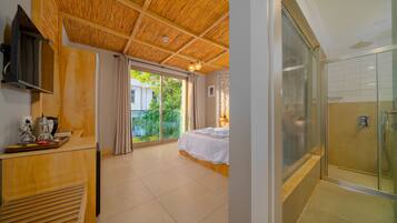 Habitación de hotel con techo de madera, cama, escritorio con cafetera, ducha con puertas de cristal y vistas a zonas verdes.