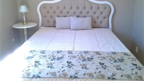 Un dormitorio con cabecera capitonada, mesita de noche redonda con lámpara y colcha estampada.