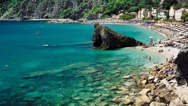 A coastal town with a rocky beach, clear turquoise waters, and buildings along the shore. - Dreamcatchers (La Spezia)