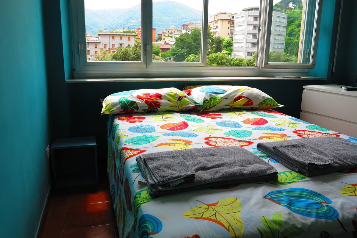 A bed with colorful leaf-patterned bedding, a window with a view of buildings and mountains, and a small bedside table.