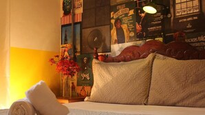 A hotel room with a bed, pillows, a rolled towel, a wall with posters, and a small table with a vase of flowers. - Mambembe Hostel - Ilha Grande (Angra dos Reis)