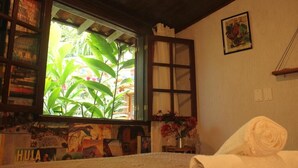 A room with a large window, a bed, a rolled-up towel, a small table with a vase of flowers, and a wall with a framed picture. - Mambembe Hostel - Ilha Grande (Angra dos Reis)