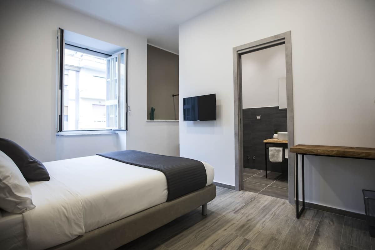 Quarto de hotel moderno com uma cama, uma TV fixada na parede, uma mesinha e uma porta para o banheiro.