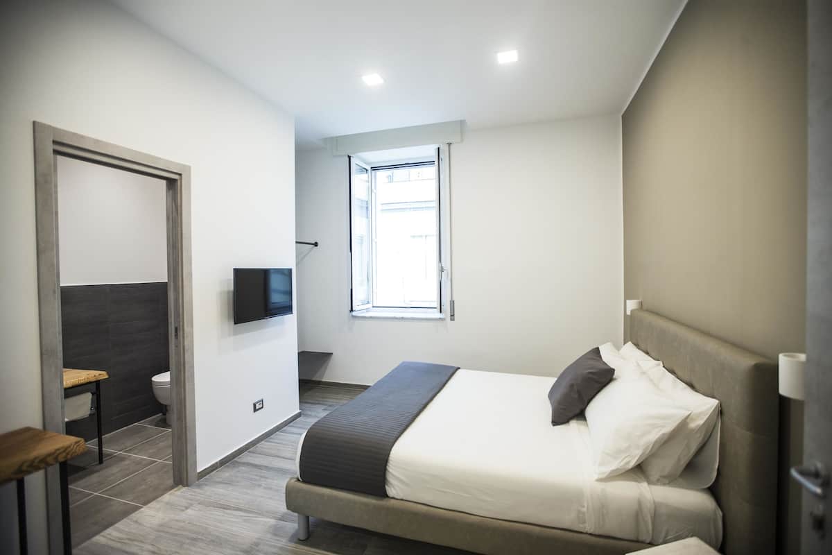 Quarto moderno com cama, televisão fixada na parede e janela que oferece luz natural.