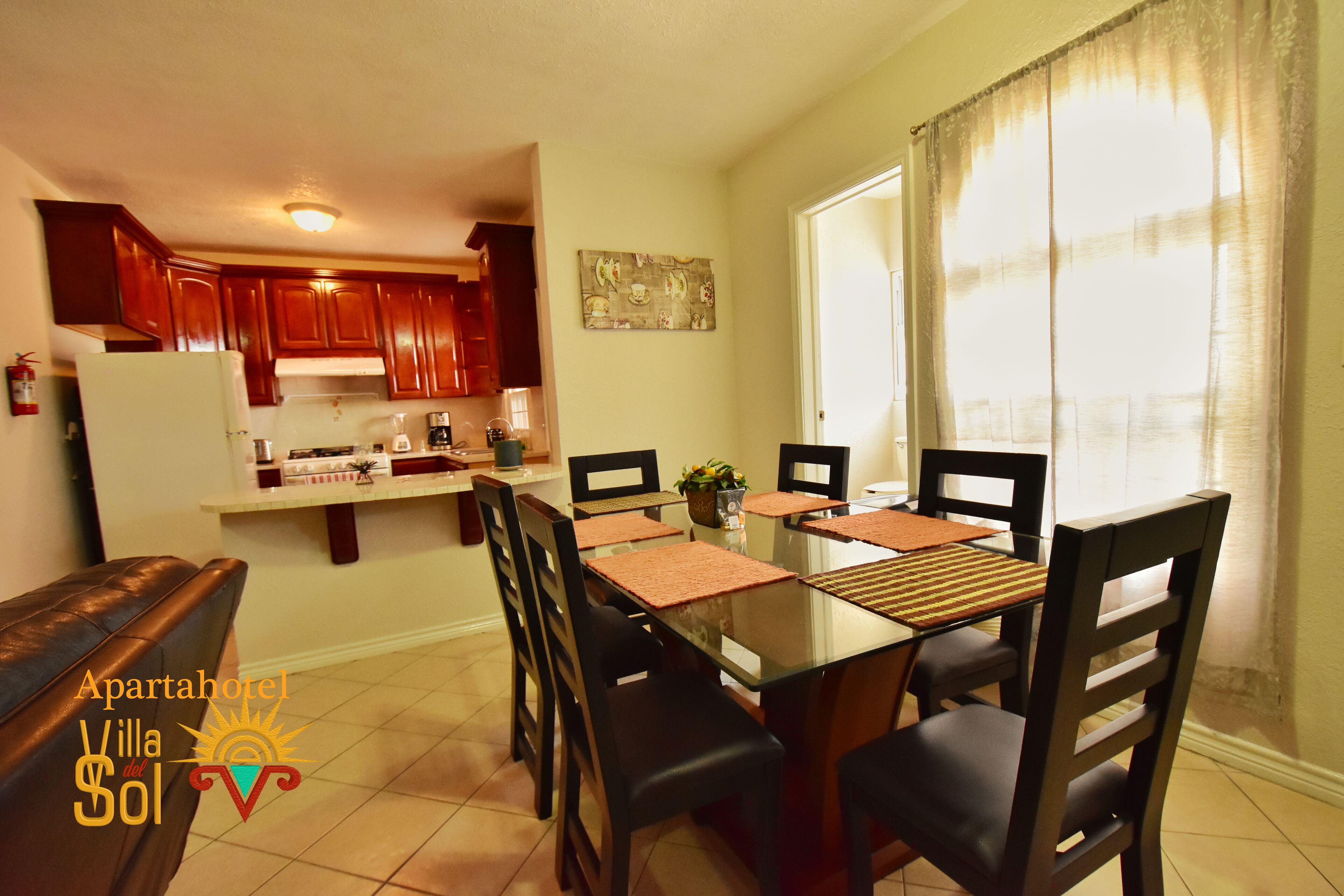 A dining area with a glass table and chairs, wooden cabinets, and a kitchen in the background.
