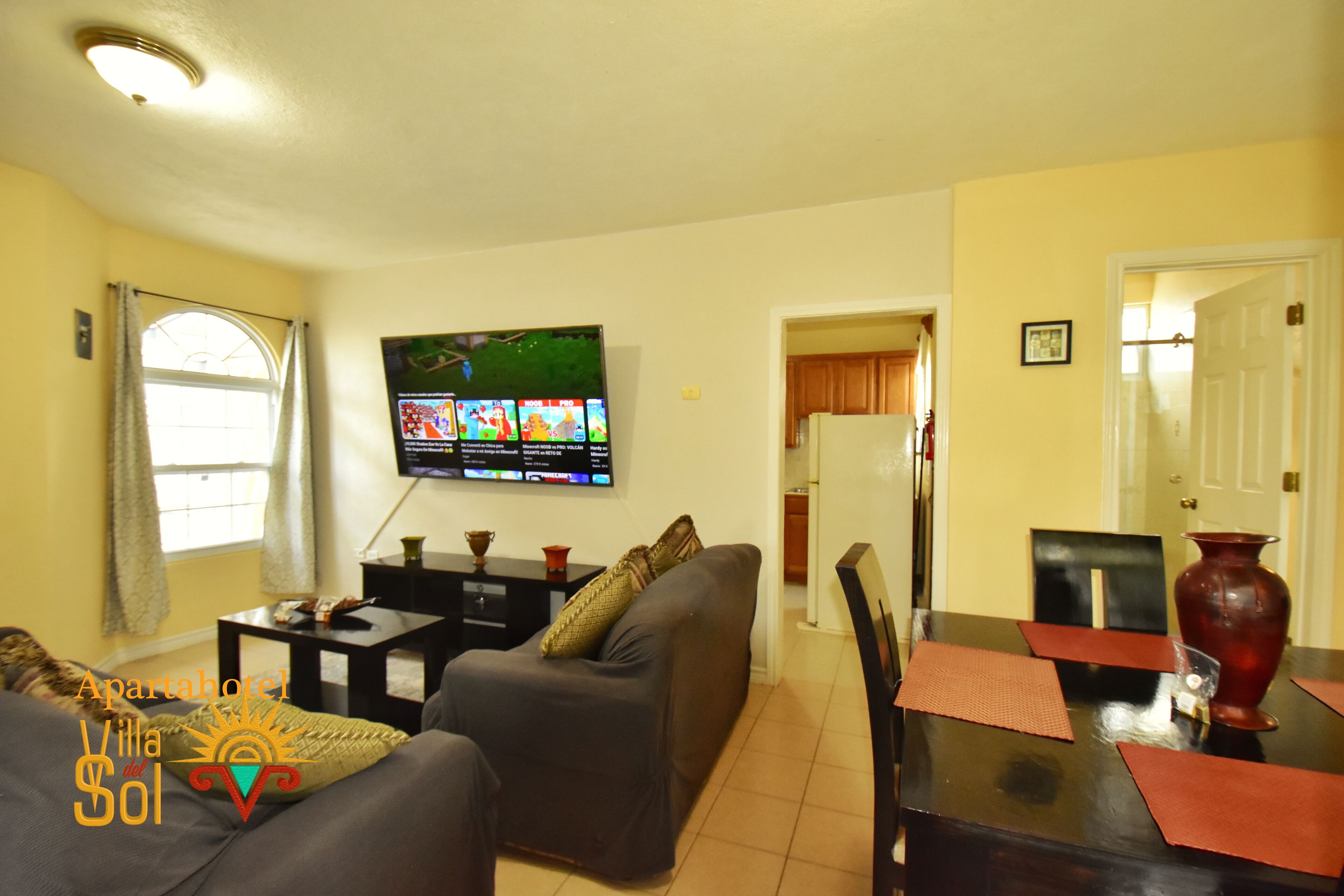 A living room with a sofa, a TV mounted on the wall, a dining table with chairs, and a kitchen area in the background.