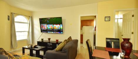 A living room with a sofa, a TV mounted on the wall, a dining table with chairs, and a kitchen area in the background.