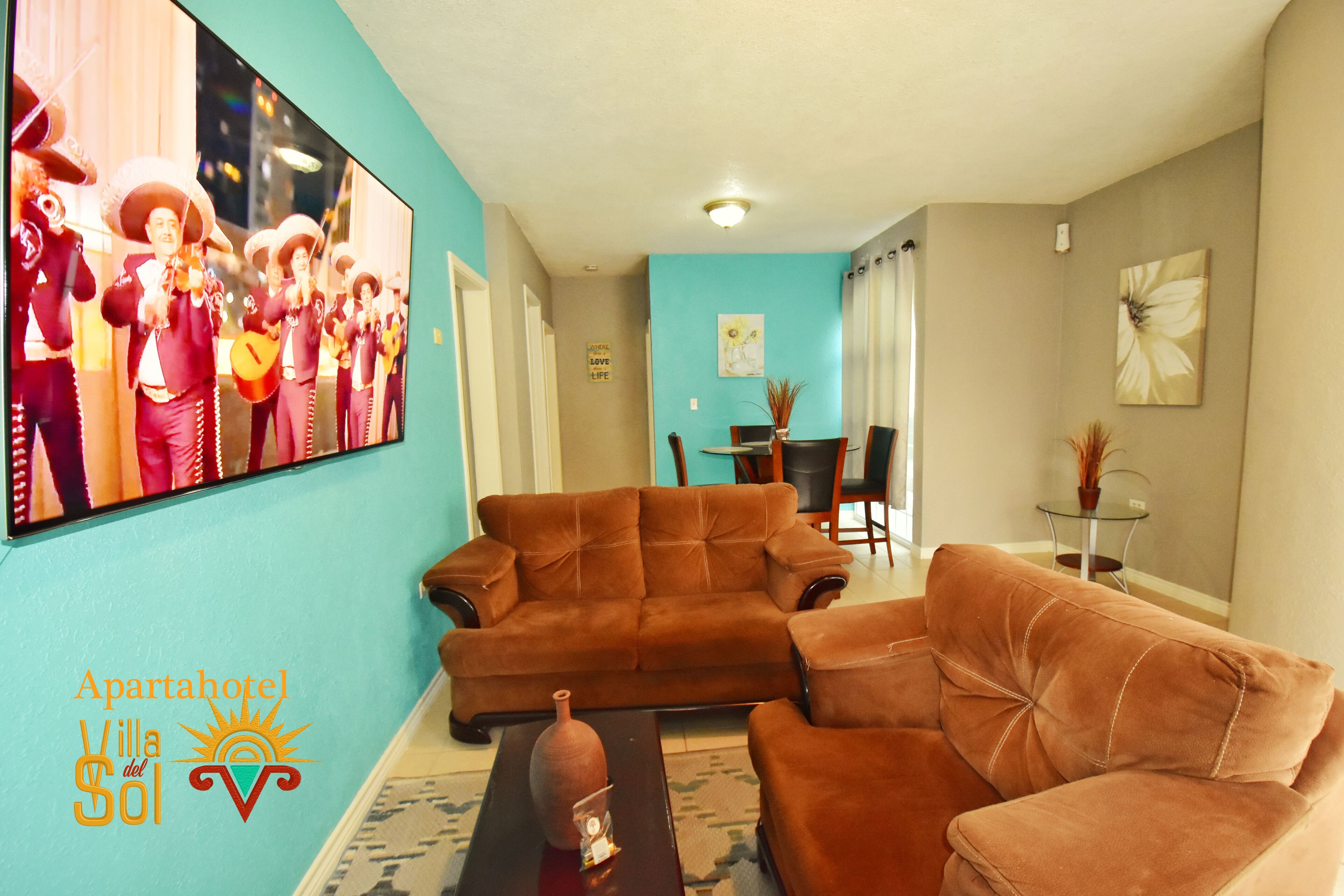 A living room with a brown sofa, a coffee table, and a painting of musicians. There is a dining area with a table and chairs in the background.