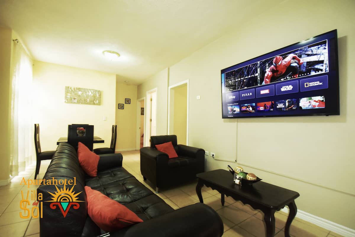 A living room with a black leather sofa, a black armchair, and a coffee table. A flat-screen TV mounted on the wall displays multiple channels.