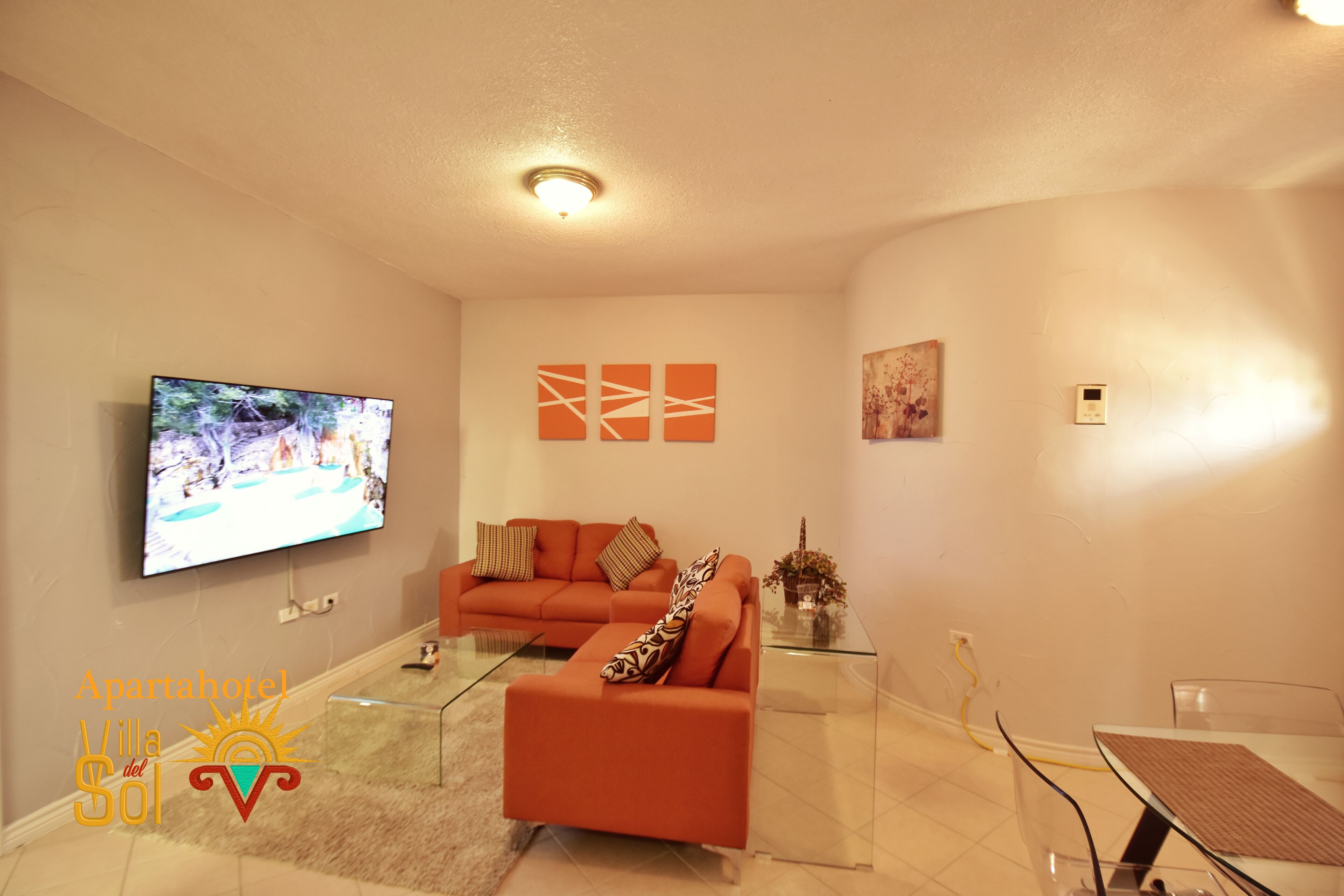 Una sala de estar moderna con televisor de pantalla plana, sofá naranja, mesa de centro de vidrio y una pequeña mesa de comedor.