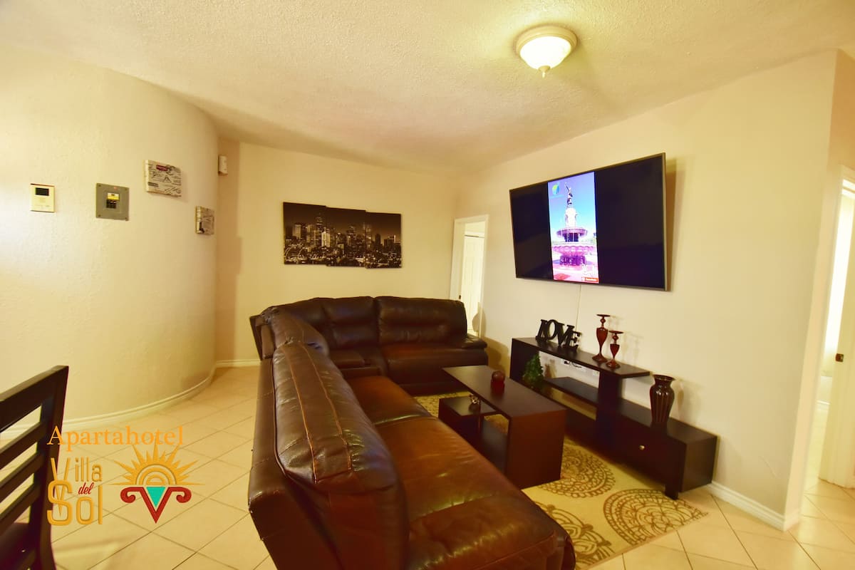 A living room with a large L-shaped leather sofa, a flat-screen TV mounted on the wall, and a small table with decorative items.