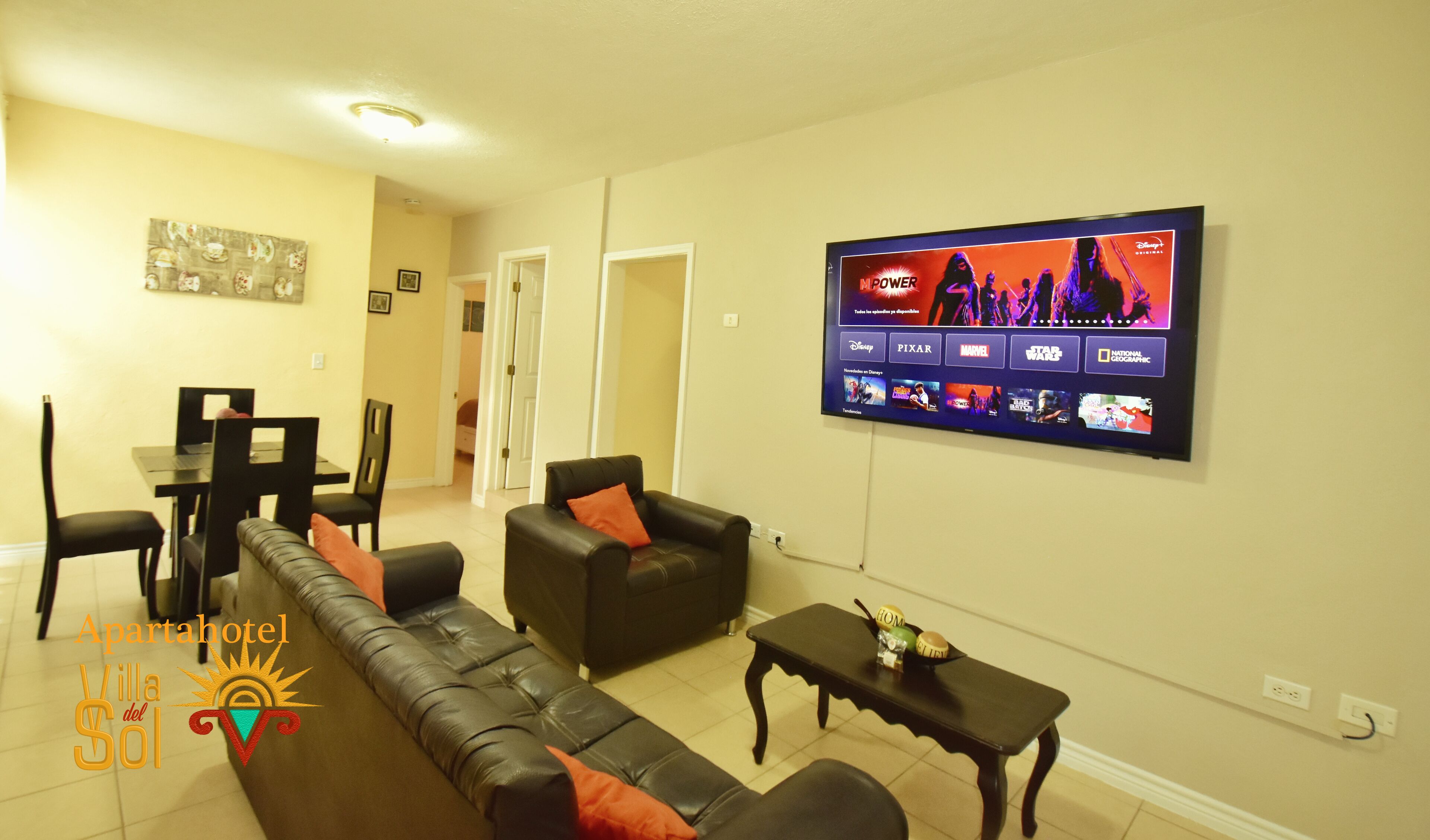 A living room with a flat-screen TV displaying various movie logos, a sofa, a coffee table, and a dining area with chairs and a table.