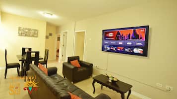 A living room with a flat-screen TV displaying various movie logos, a sofa, a coffee table, and a dining area with chairs and a table.