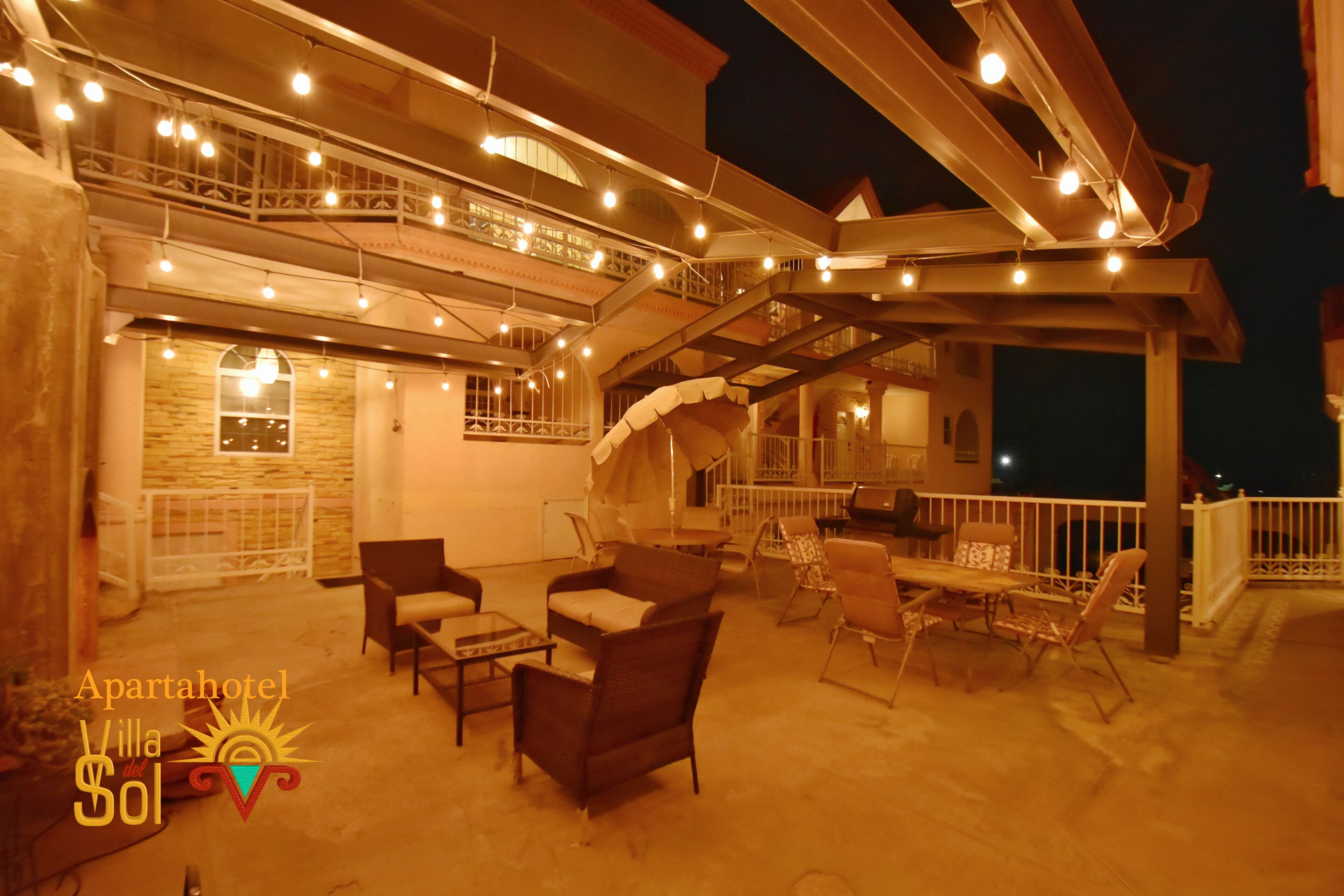 An outdoor patio area with multiple seating arrangements, including chairs and tables, under a covered structure with string lights.