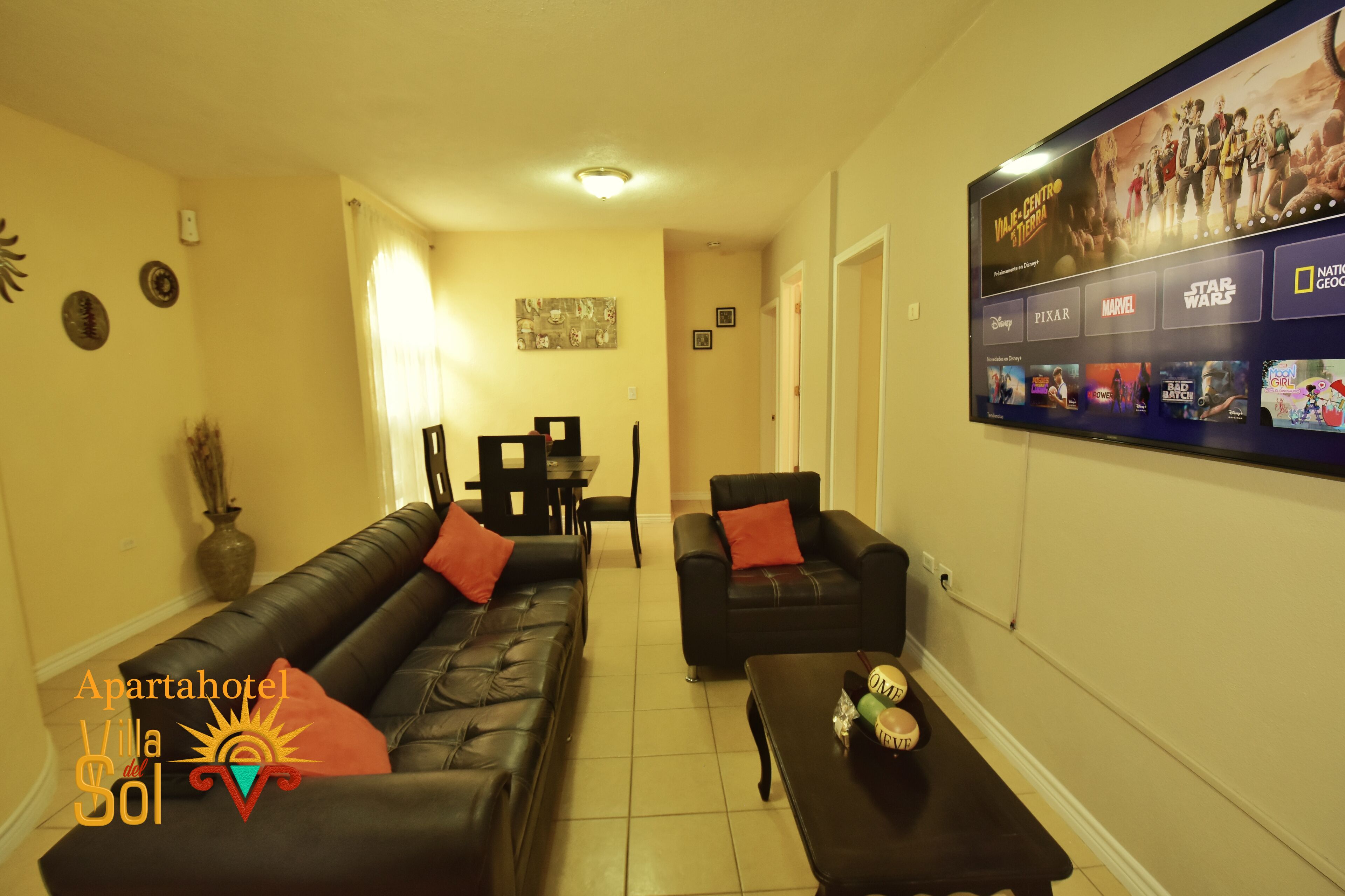 Una sala de estar con un sofá de cuero negro, un sillón negro y una mesa de centro. Hay un televisor montado en la pared mostrando opciones de películas.