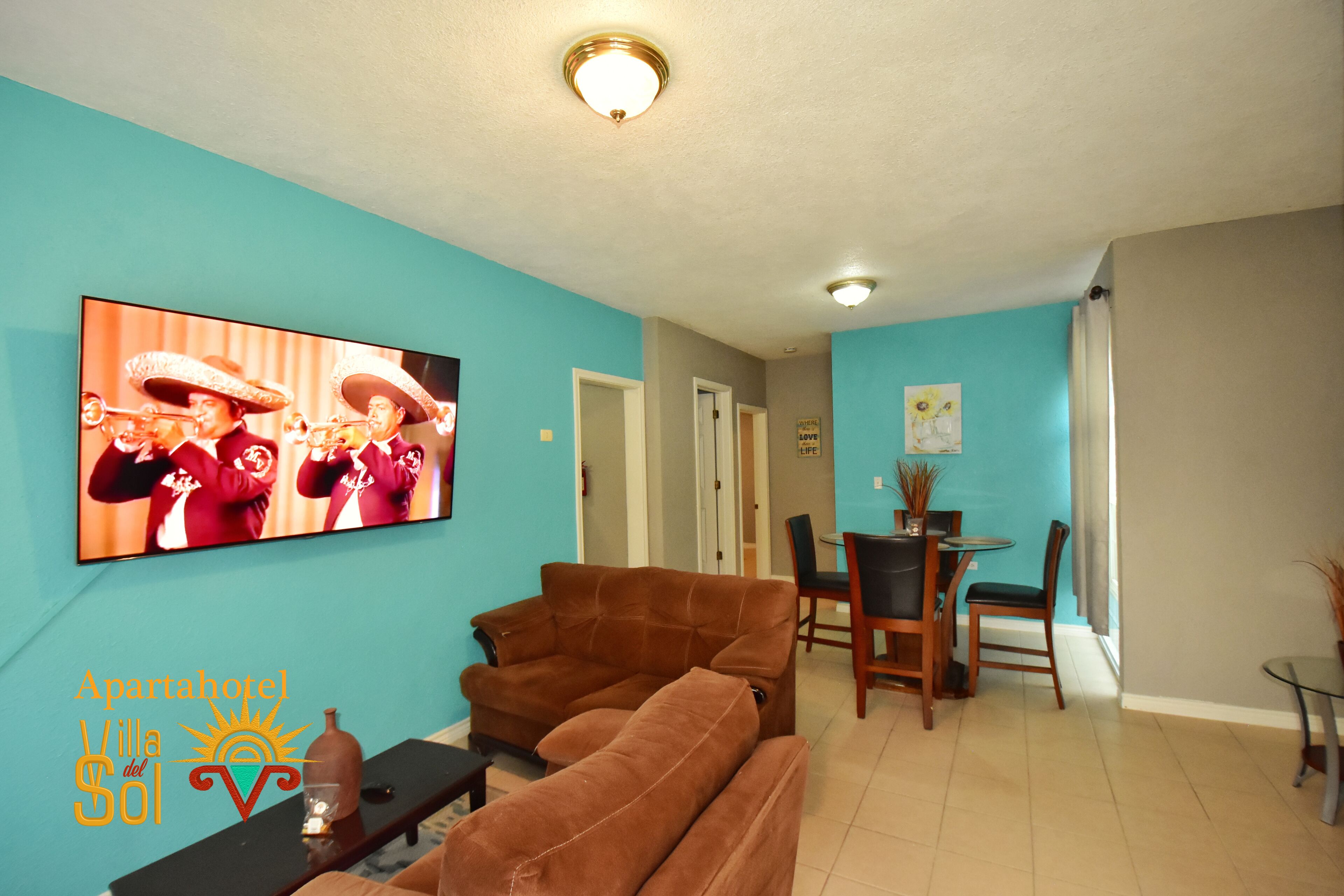 A living room with a brown sofa, a TV displaying a band, and a dining area with chairs and a table.