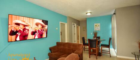 A living room with a brown sofa, a TV displaying a band, and a dining area with chairs and a table.
