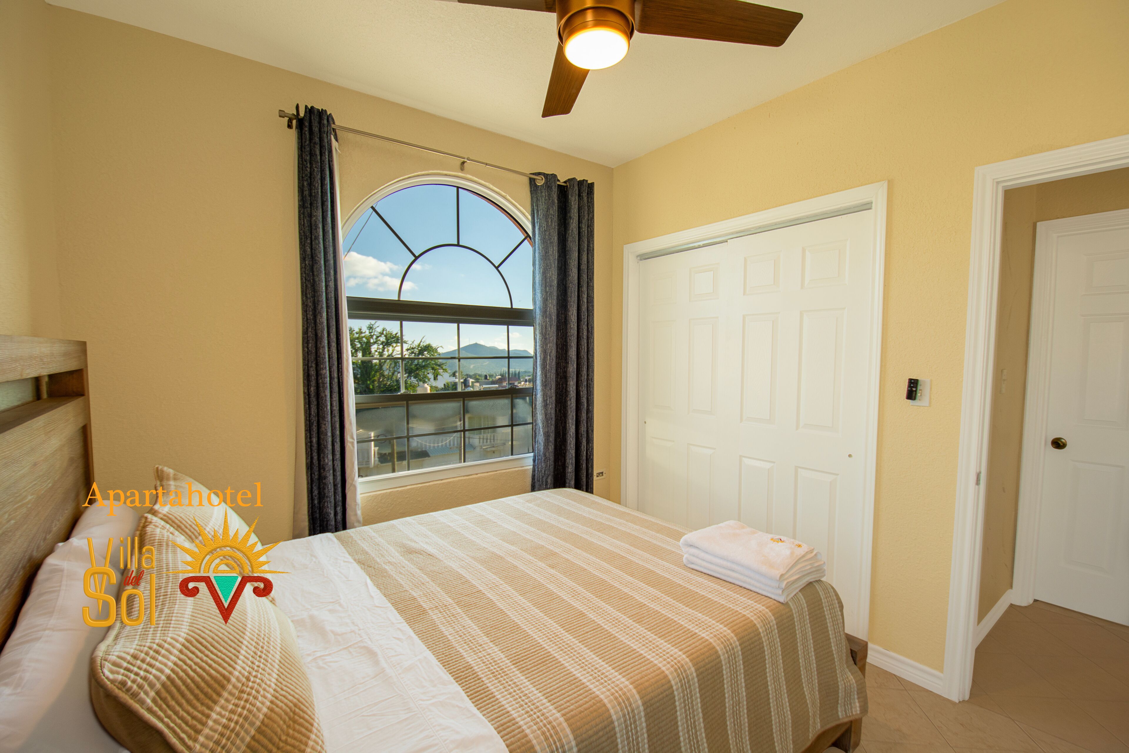 A bedroom with a bed, a window with curtains, a ceiling fan, and a door.