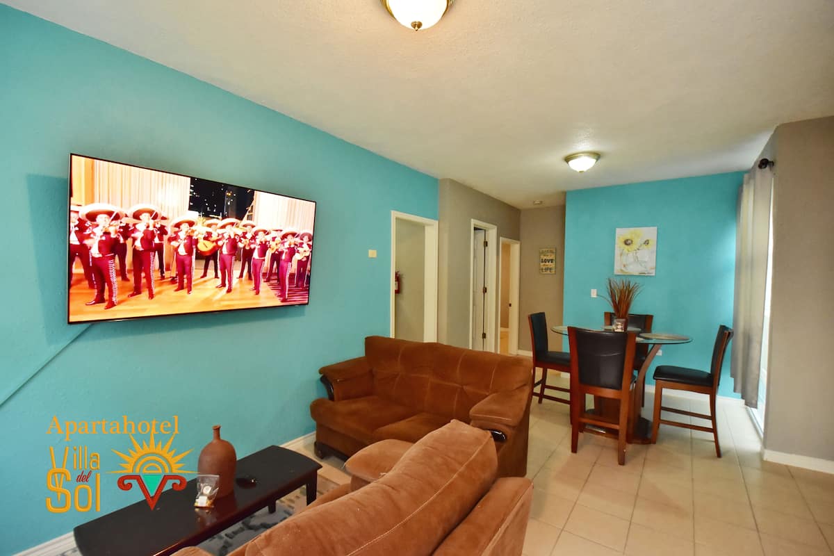 A living room with a brown sofa, a TV mounted on the wall, and a dining area with a table and chairs.