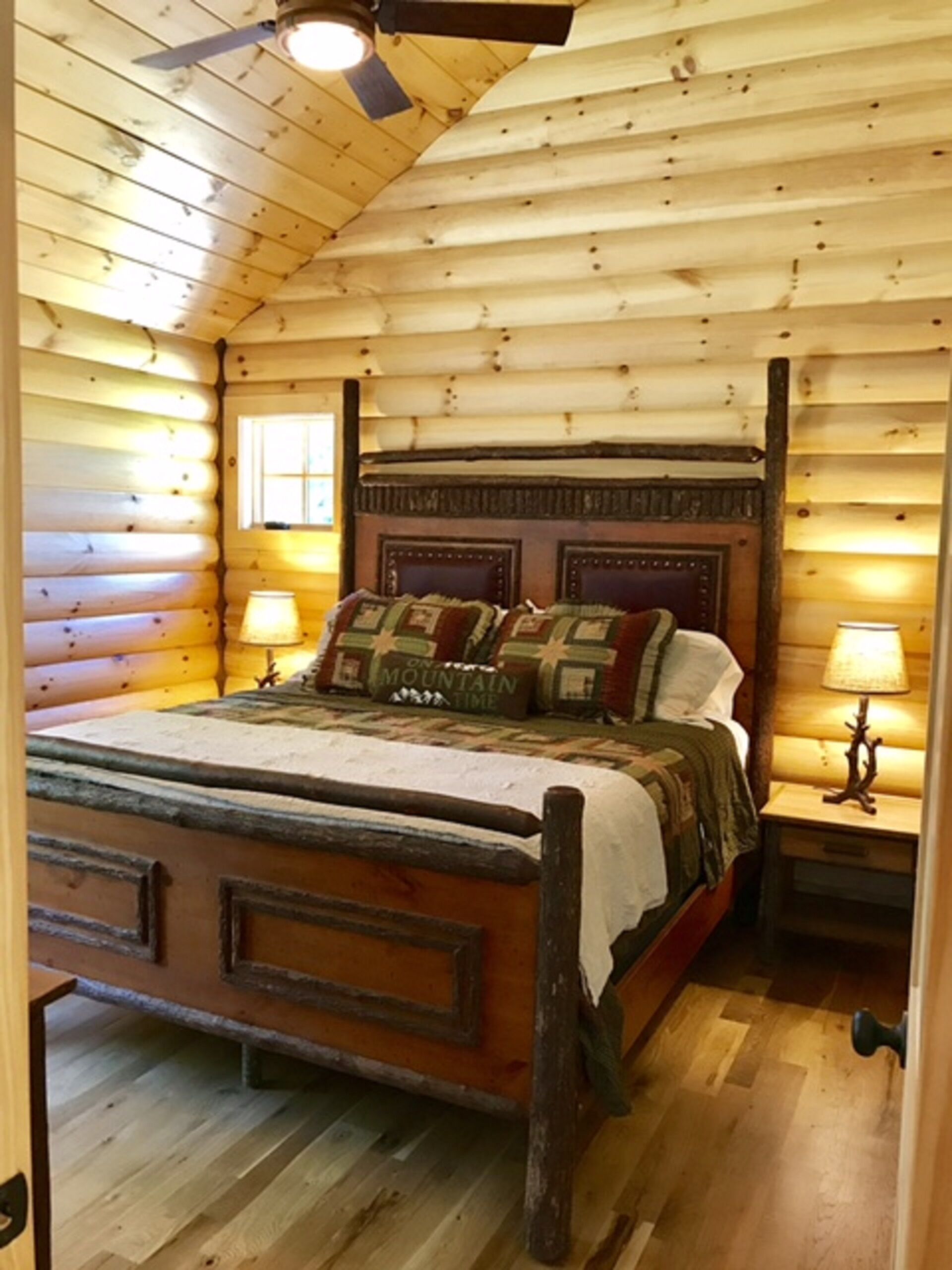 Een slaapkamer in een blokhut met een houten bed, twee bedlampjes en een plafondventilator.