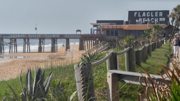 Een strand met een houten omheining, een steiger en een gebouw met de tekst 'FLAGLER BEACH'.
