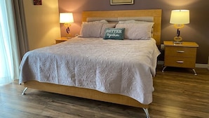 Quarto de hotel com uma cama grande, duas mesas de cabeceira com luminárias, um quadro na parede e piso de madeira.