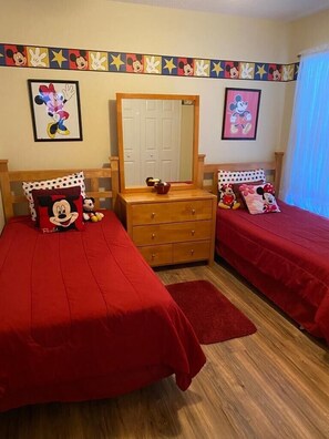 A room with two beds, each with a Mickey Mouse themed pillow, a wooden dresser, and a mirror.