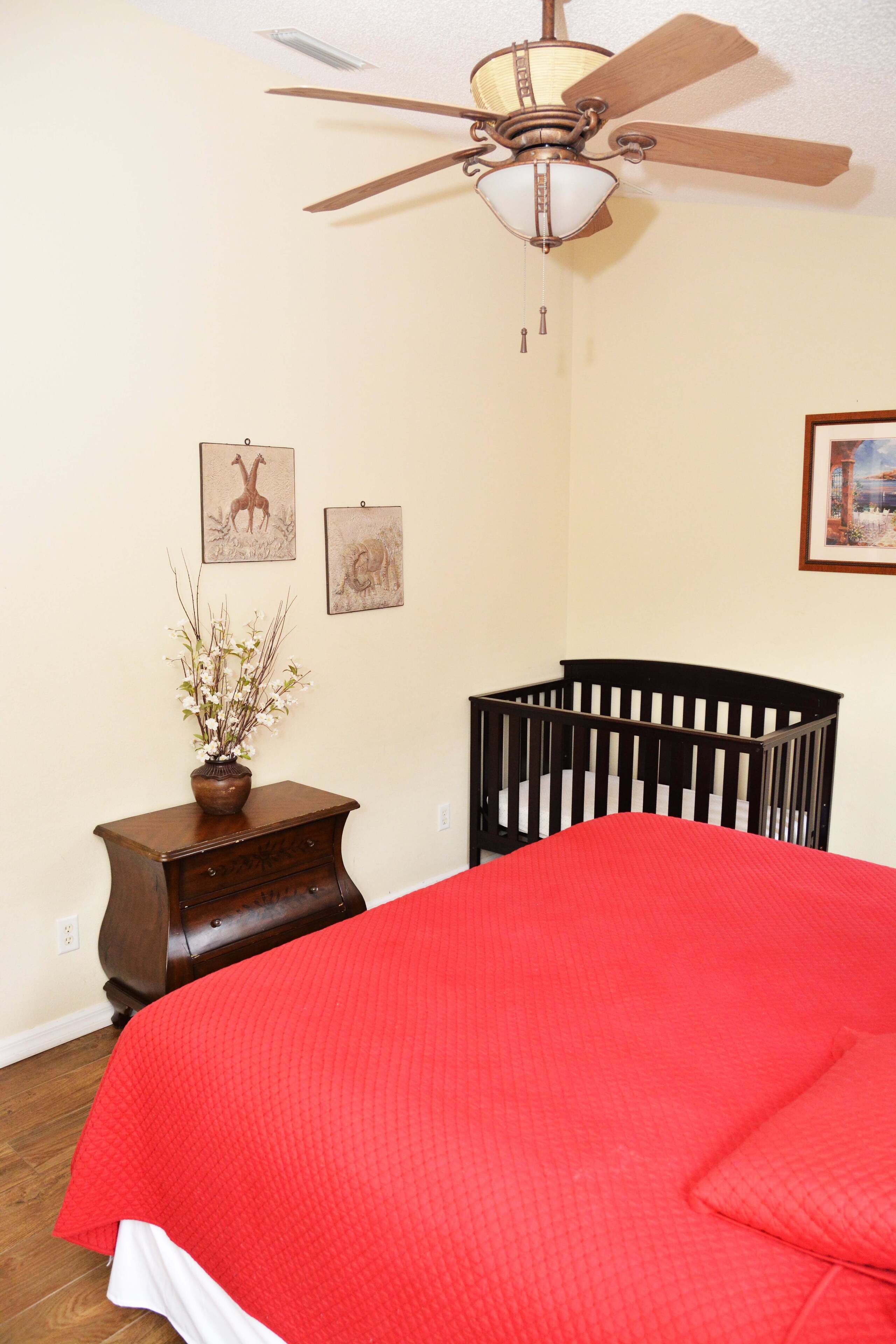 A bedroom with a red bedspread, a wooden crib, a ceiling fan, and wall art.
