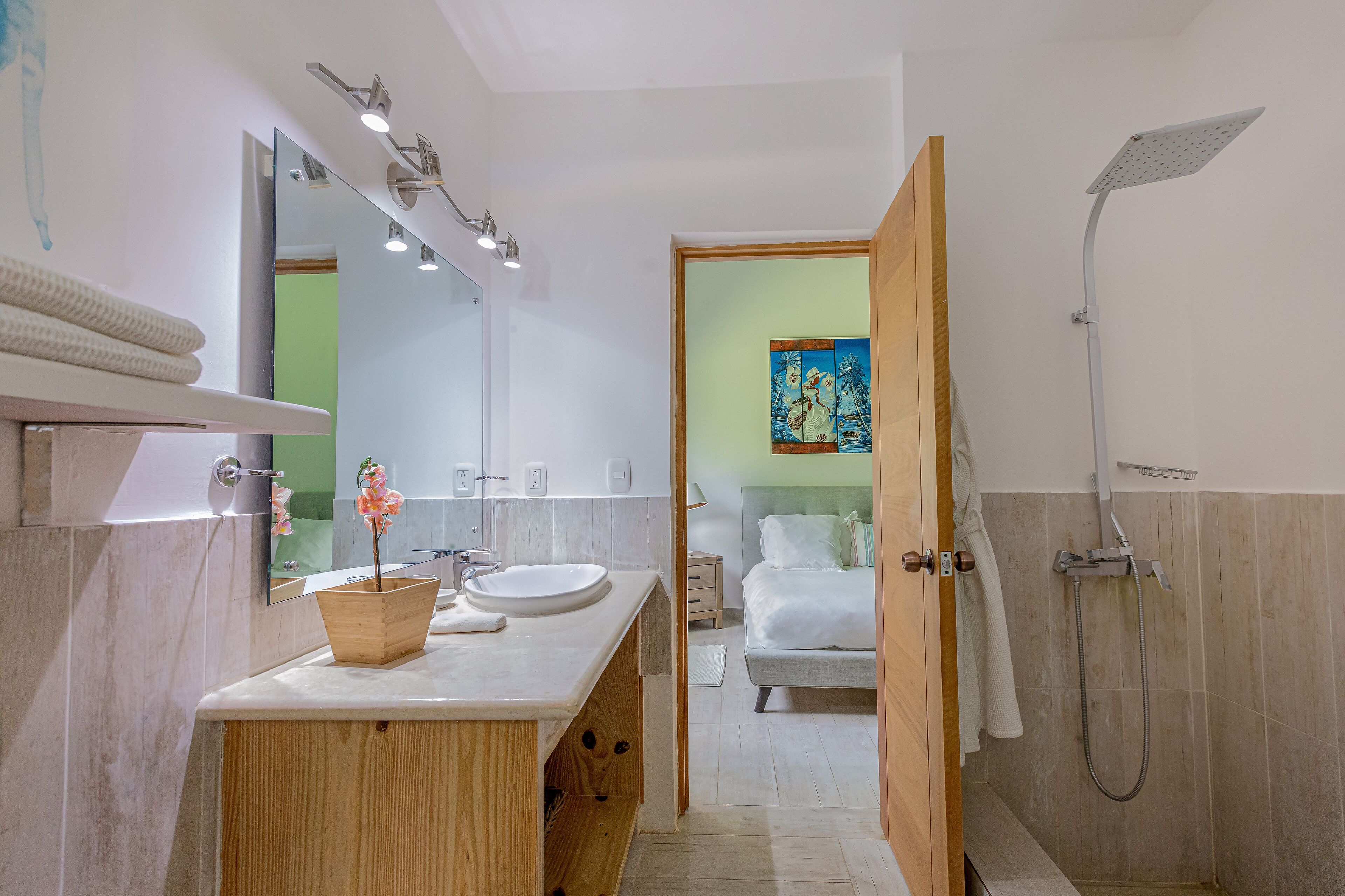 A modern bathroom with a wooden vanity, a large mirror, a shower, and a bed visible through an open door.