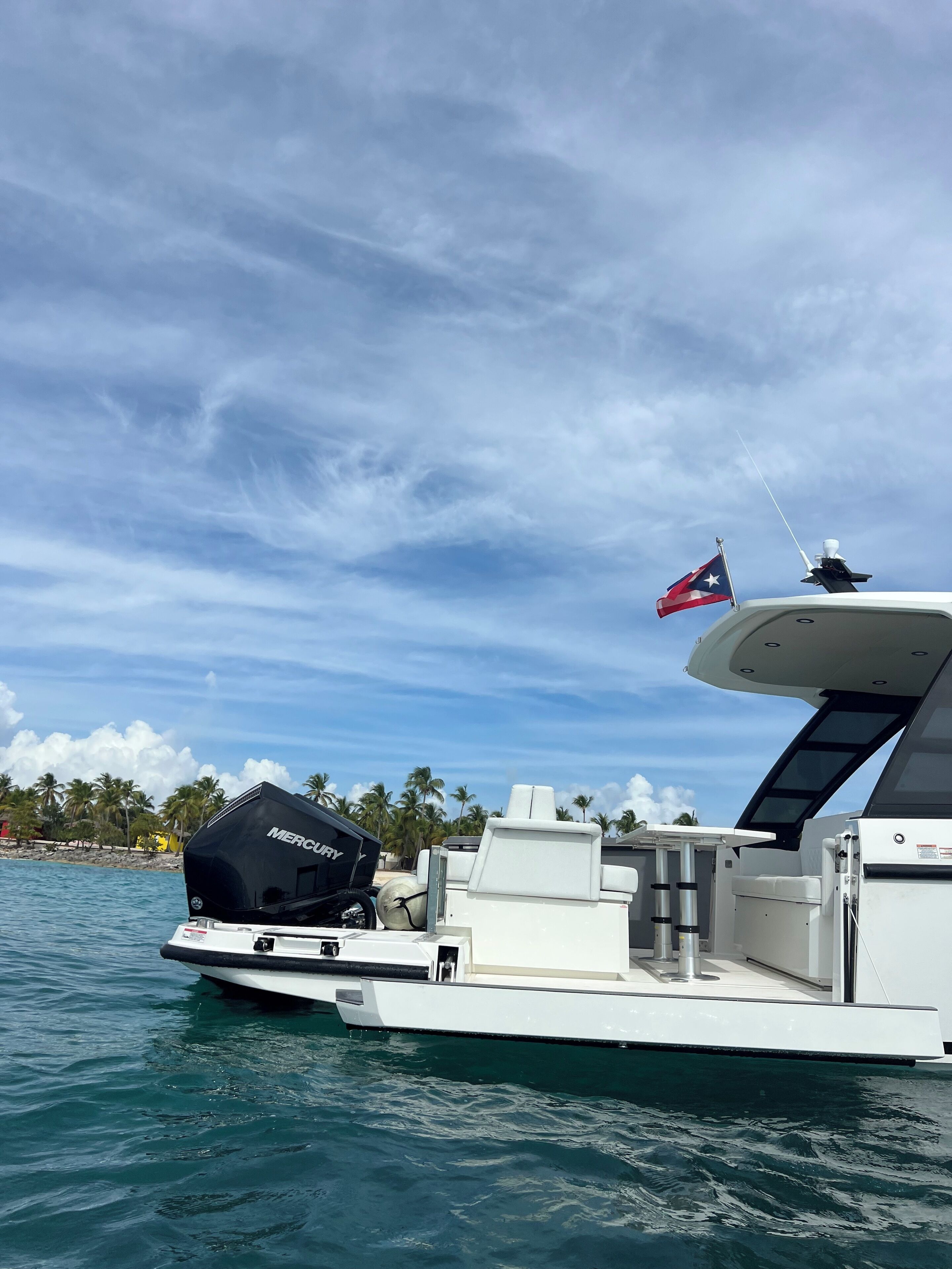 Un yate blanco con motor a la vista, una bandera y una vista despejada del océano y palmeras al fondo.