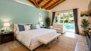 Une chambre dotée d’un grand lit, de tables de chevet, d’un banc, d’une chaise, et offrant une vue sur une piscine, ainsi que d’un ventilateur de plafond.