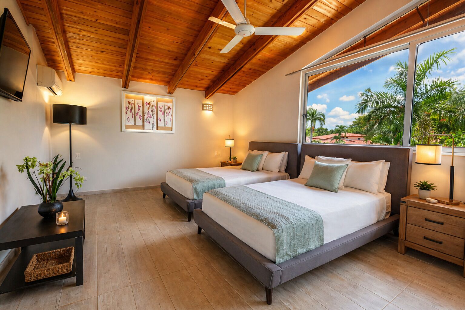 A double room with two beds, a ceiling fan, a large window with a view, and a small table with a lamp.