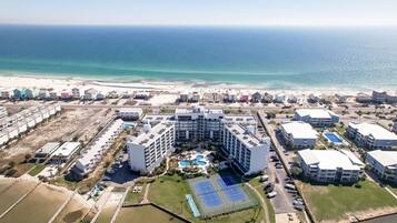 An aerial view of a coastal hotel complex with multiple buildings, a central swimming pool, tennis courts, and a beach in the background.