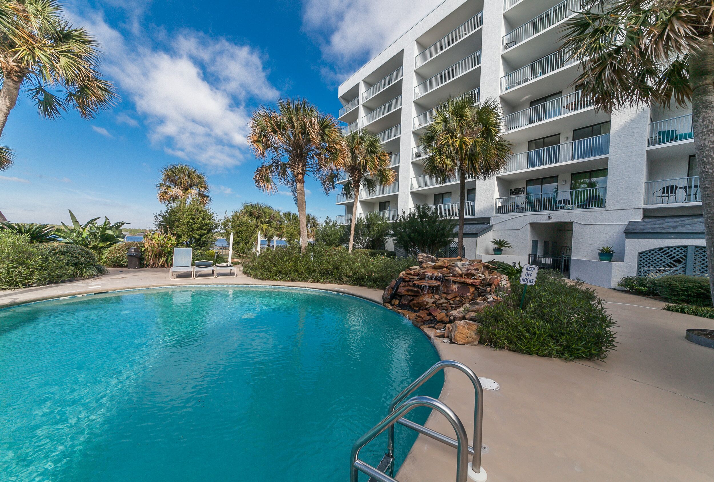 A hotel with a swimming pool, palm trees, and a stone waterfall feature.