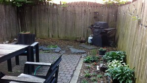 Un jardin avec une table et des chaises en bois, un barbecue et un espace planté de différentes espèces végétales et de fleurs.