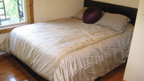 Un lit bien fait, recouvert d’une couvre-lit beige agrémentée d’une bordure décorative, une fenêtre donnant sur des arbres et un plancher en bois.