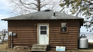 A small wooden cabin with a white door, a small window, and a chimney. There is a sign for Freshwater Vacation Rentals and a propane tank nearby.
