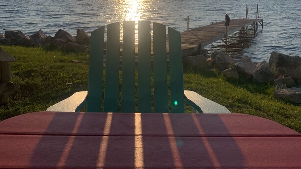A wooden deck with a red table and a green chair facing a body of water at sunset.