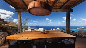 Ein Holztisch mit Stühlen unter einer Holzpergola, mit Blick auf die Küste, Felsen und das Meer.