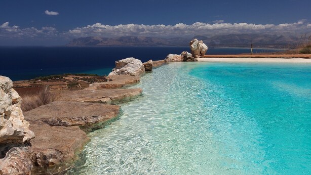 Ein klares, blaues Schwimmbecken mit steiniger Kante, umgeben von einer Küstenlandschaft und Bergen im Hintergrund.