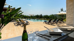 Ein Poolbereich mit Liegestühlen, einem Tisch mit Weingläsern und einer Flasche, sowie Blick auf Pool und Grünflächen.