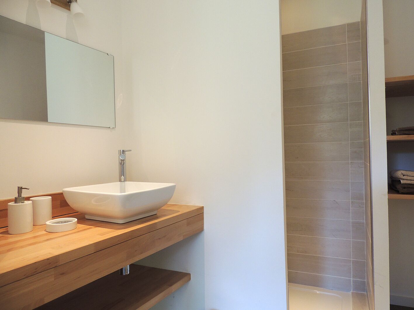 Un baño moderno con un lavamanos blanco de diseño, un mueble de tocador de madera, un espejo y una zona de ducha con azulejos.