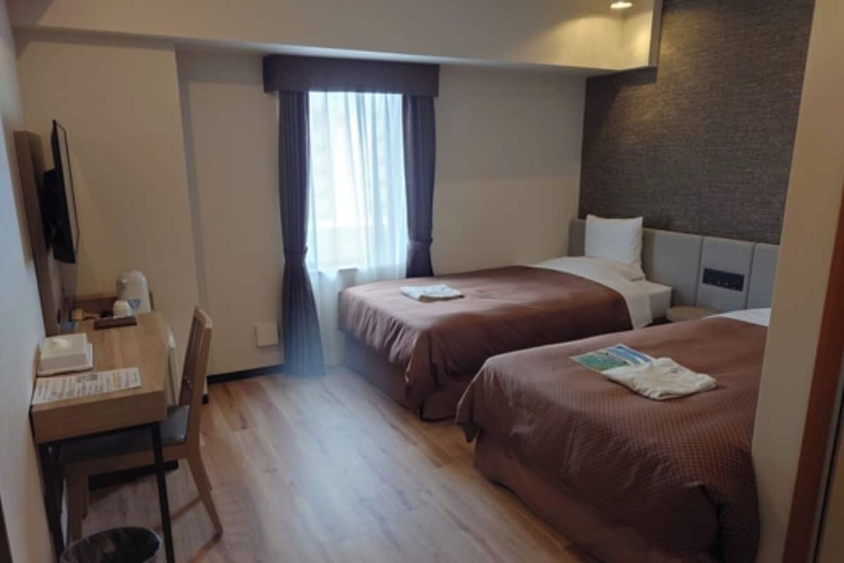 Ein Hotelzimmer mit zwei Betten, einem Schreibtisch, einem Stuhl, einer Mülltonne und einem Fenster mit Vorhängen.