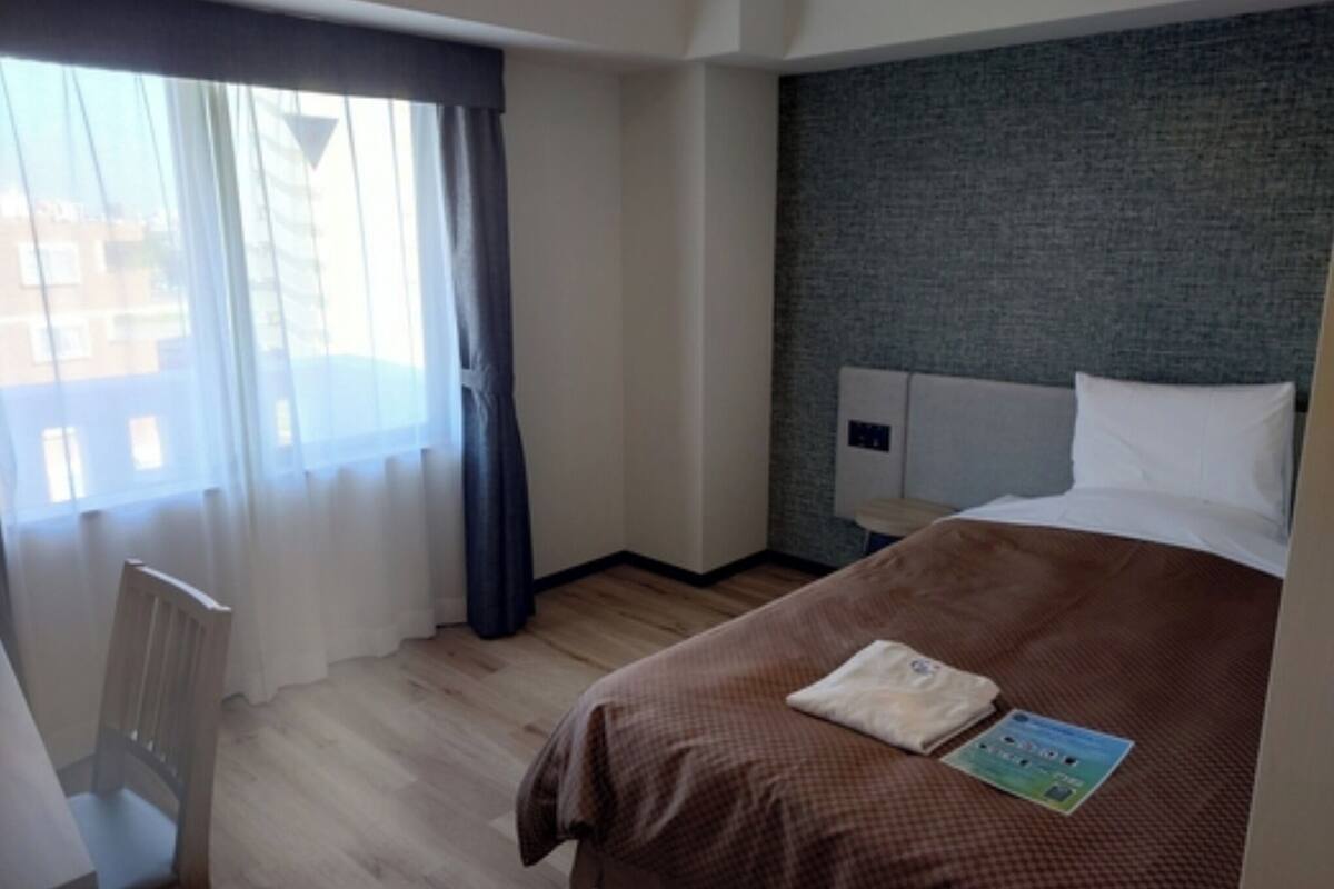 Ein Hotelzimmer mit Bett, Stuhl, einem Fenster mit Vorhängen und Blick auf Gebäude.