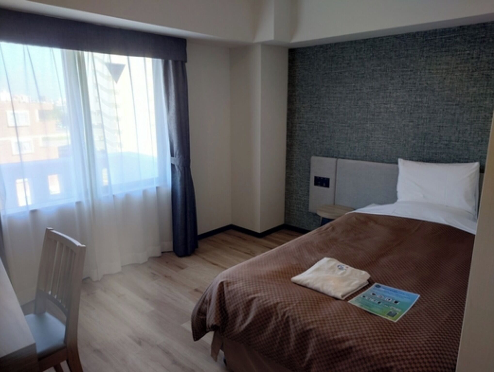 A hotel room with a bed, a chair, a window with curtains, and a view of buildings outside.